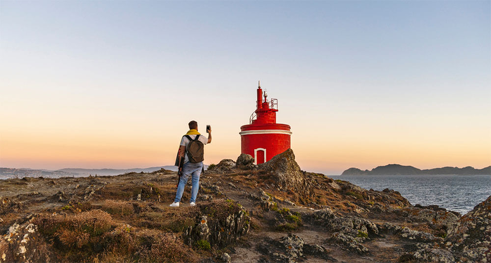 西班牙北部陡峭的欧洲大西洋沿岸的一座红色灯塔，年轻人用他的手机拍照。